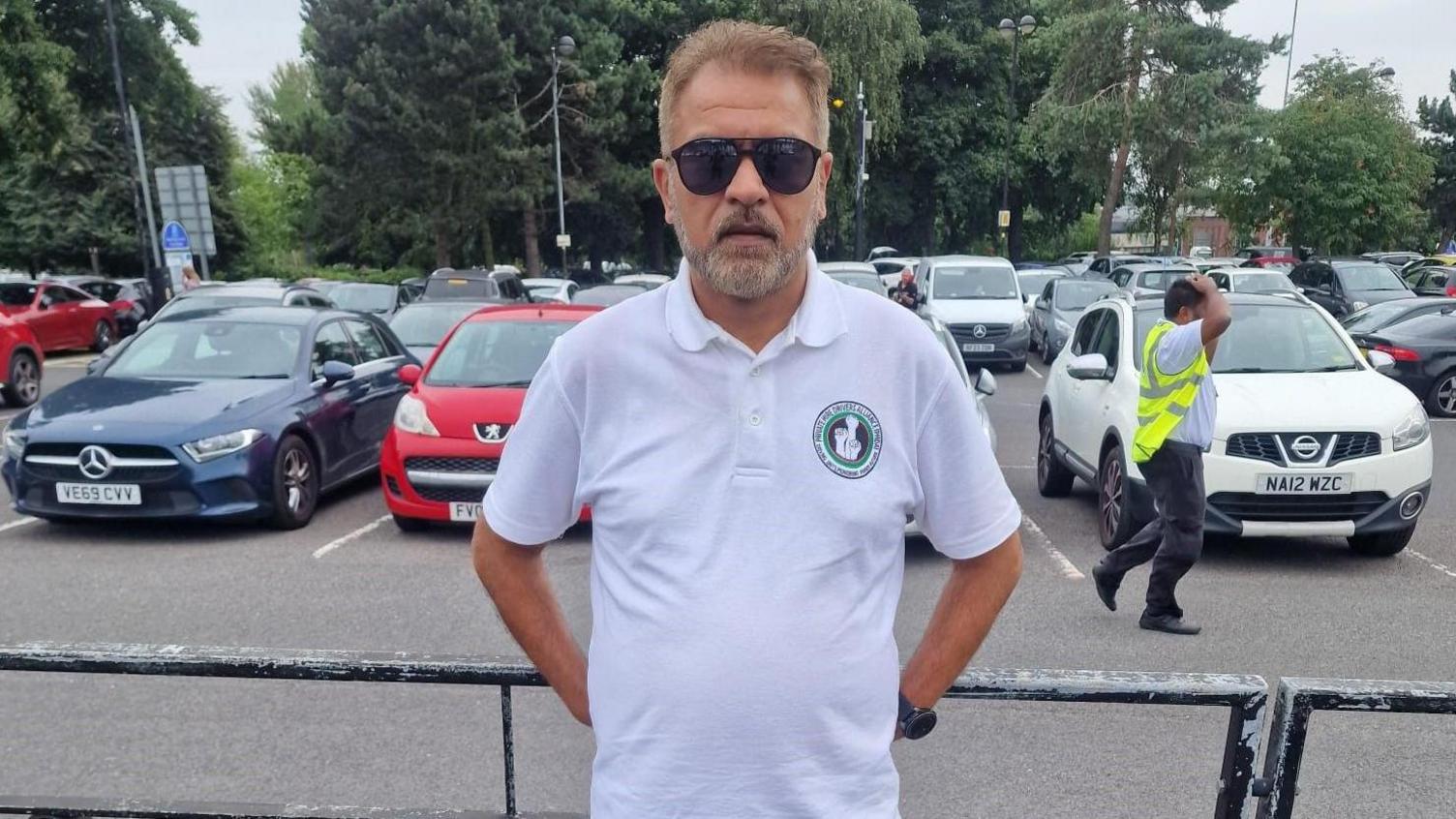 Saj Ahmed is wearing a white T-shirt and sunglasses while leaning against a railing