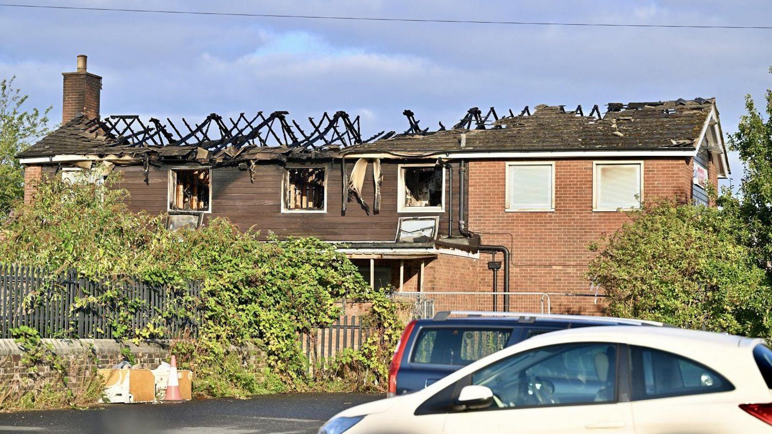 The full extent of the fire damage viewed from a nearby road. The charred woodwork, bricks and tiles are visible. The windows of the pub have also been broken and the interior rooms have been gutted.