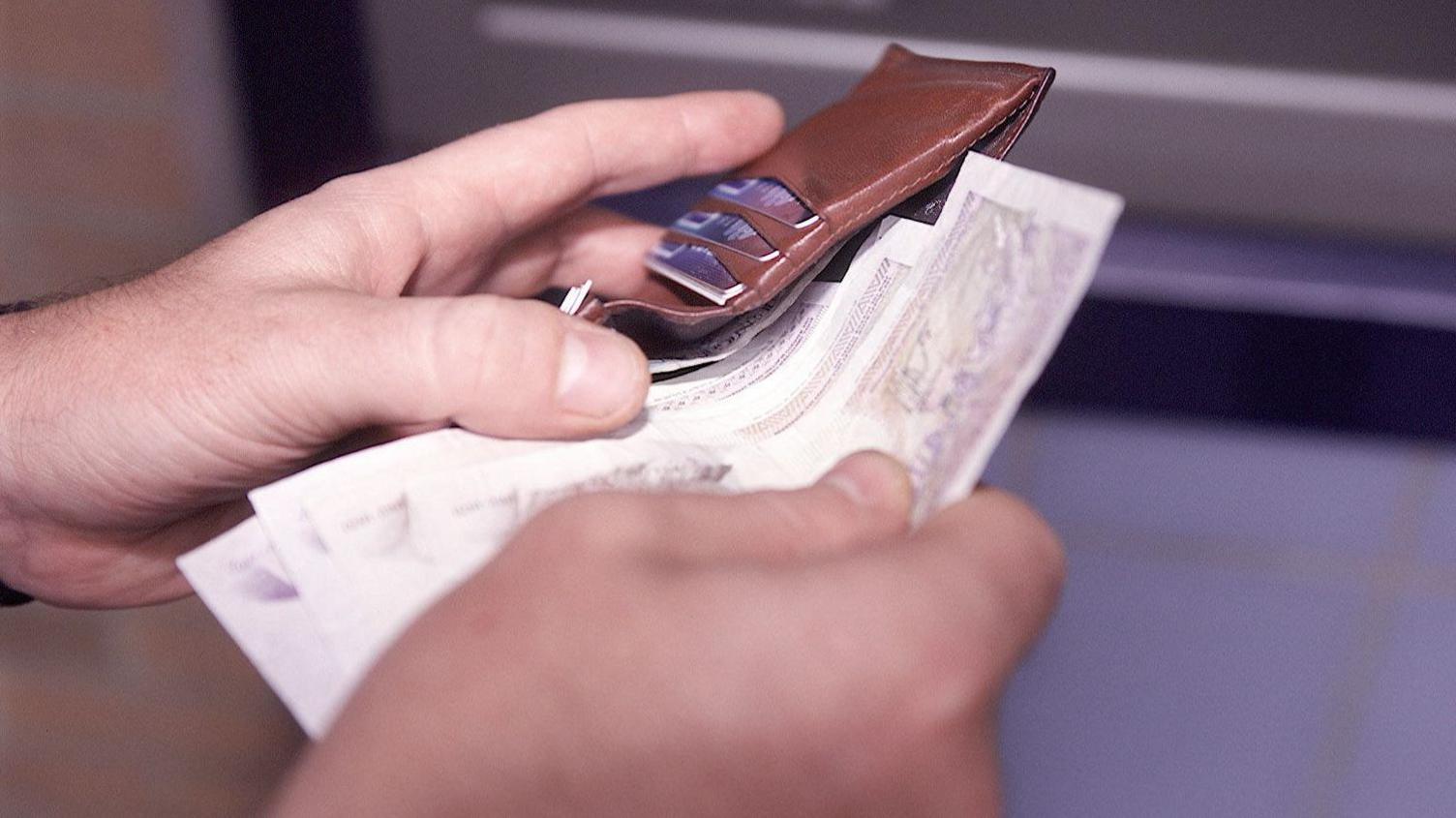 A man holds a brown leather wallet with his left hand and is taking bank notes out of it with his right hand.