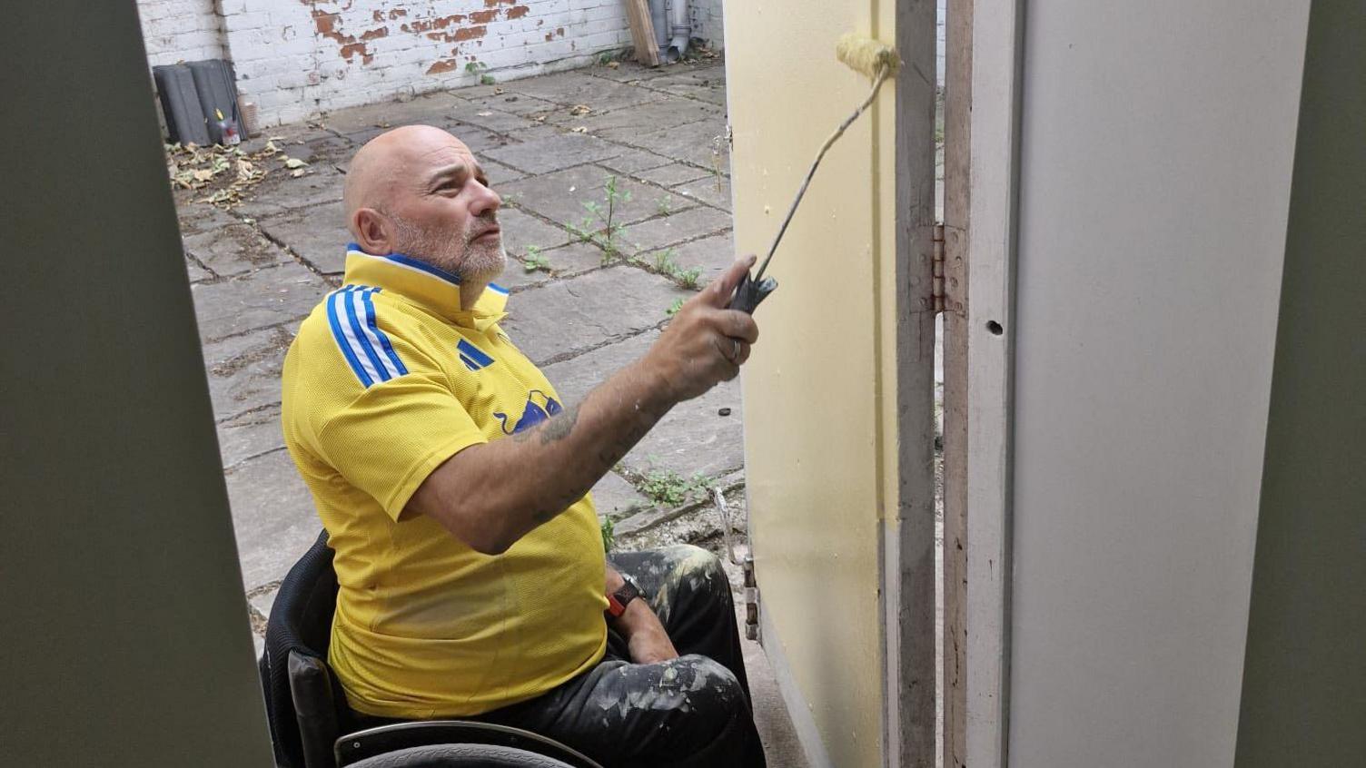 Man with yellow football shirt in a wheelchair painting a door with a roller and yellow paint.