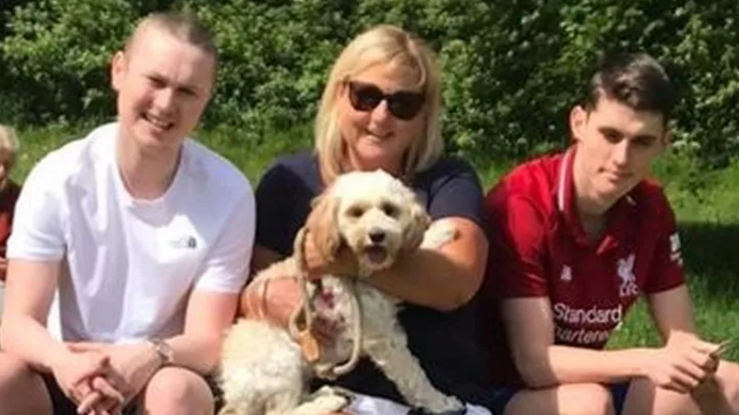 Ben with his mother Jo and brother Charlie. They are sitting down in a group on a sunny day, with Ben wearing a white T-shirt and his brother wearing a red Liverpool top. His mother is holding a small white dog