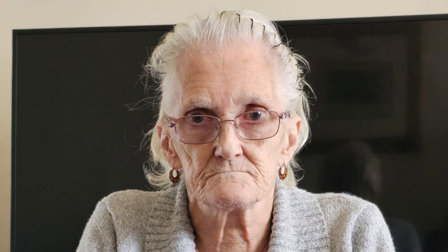 An elderly woman with shoulder length grey hair looks directly at the camera. She is wearing a grey jumper and glasses. She has an unhappy expression.