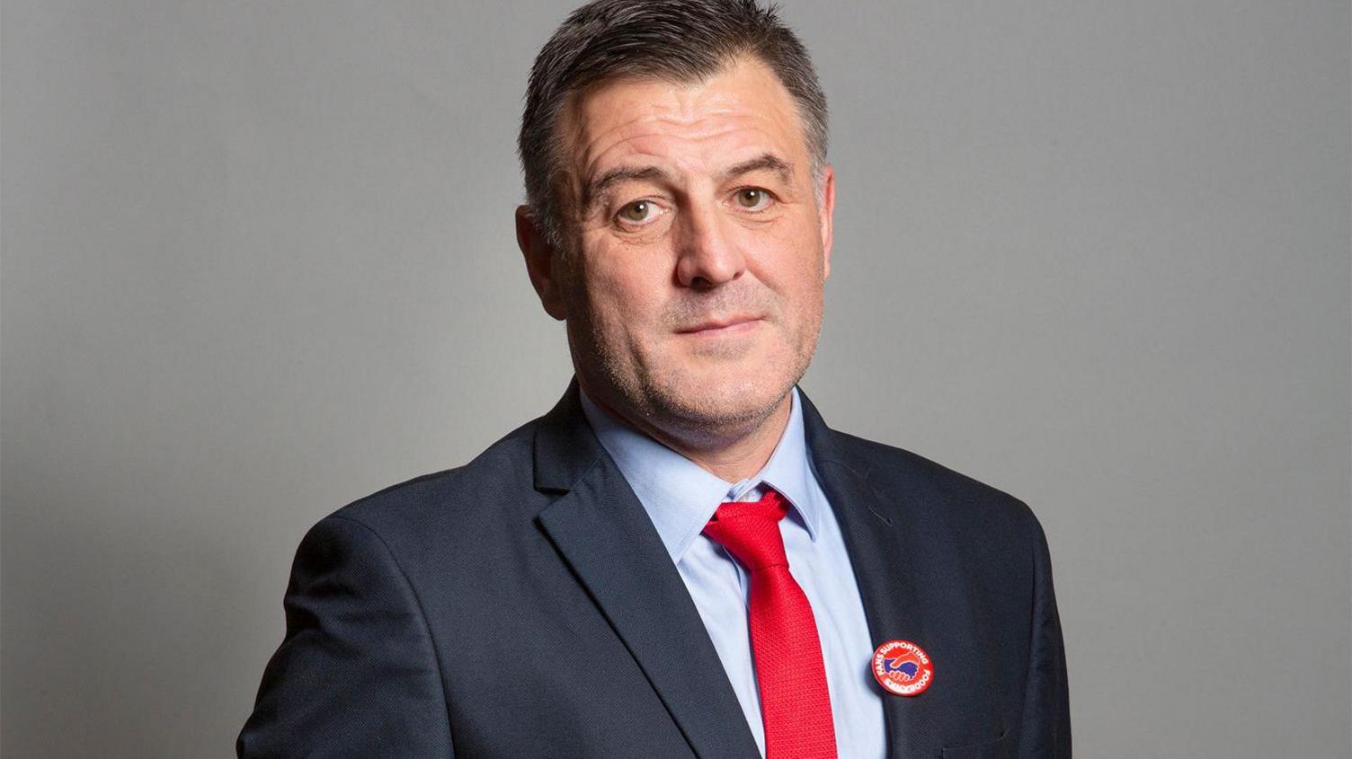 Ian Byrne, who has dark hair greying at the sides, poses for a picture wearing a navy blue suit with a red tie. 