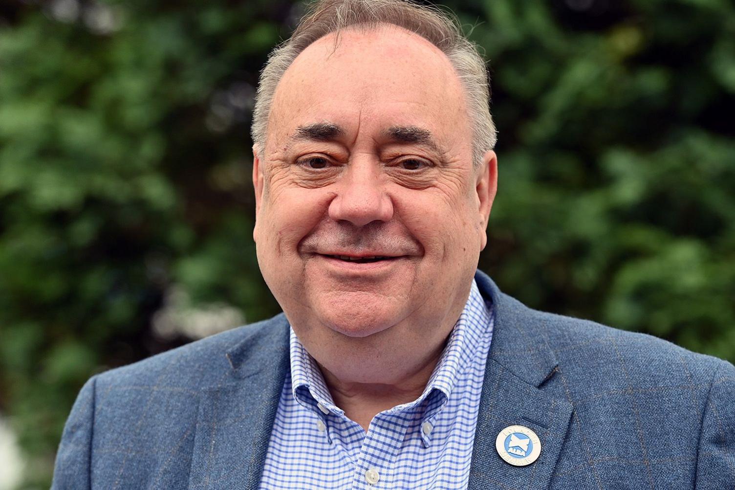 Alex Salmond is photographed outside against a blurry background of trees. He is bald and clean-shaven, with distinctively bushy eyebrows, and smiles for the camera. He wears a light blue jacket with an "Alba" badge pinned to it, and a blue-check button-down shirt.