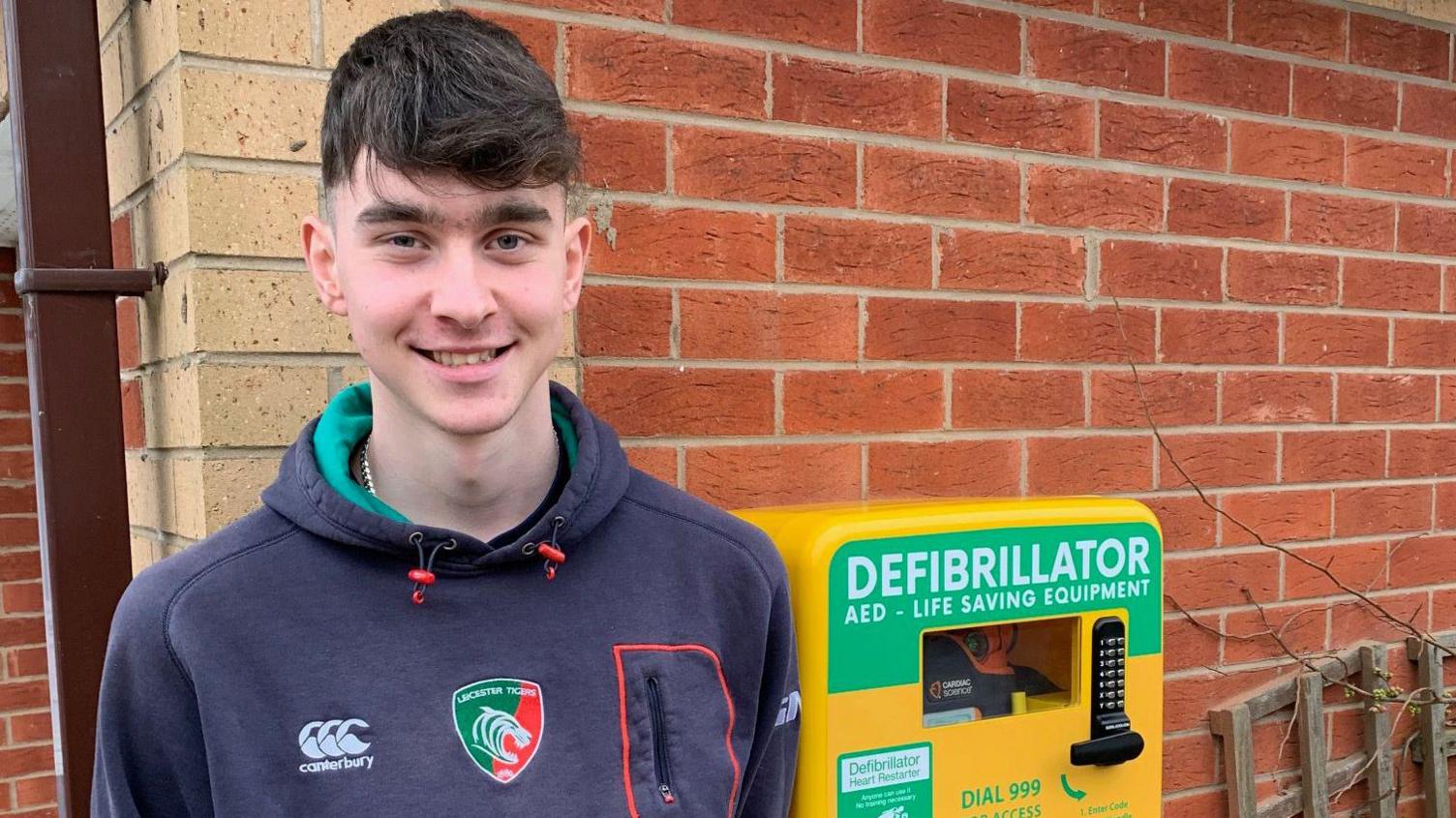 A young man in a Tigers hooded top standing next to a bright yellow defibrillator box on the side of a brick building