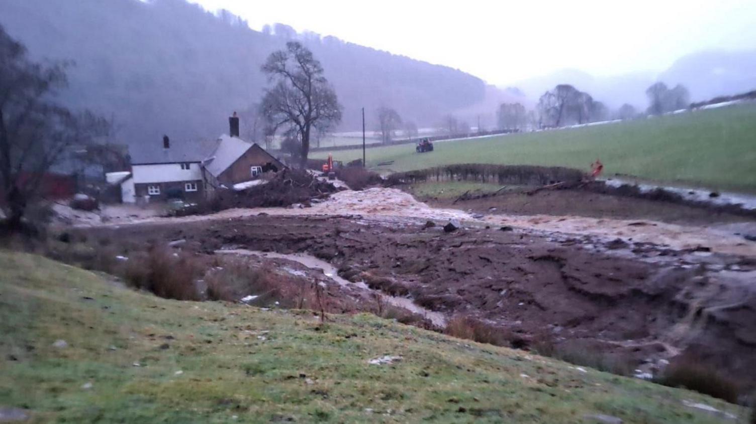 A brick farmhouse pictured from a distance with tonnes of brown silt covering the yard in front of it. A pile of branches and fallen trees can also be seen by the farmhouse.