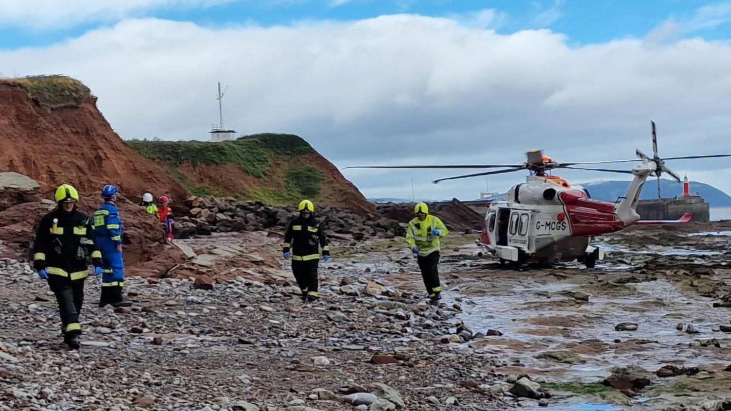 Six rescuers standing on a pebbled beach near a red and white helicopter. There are verges to the right-hand side of the image