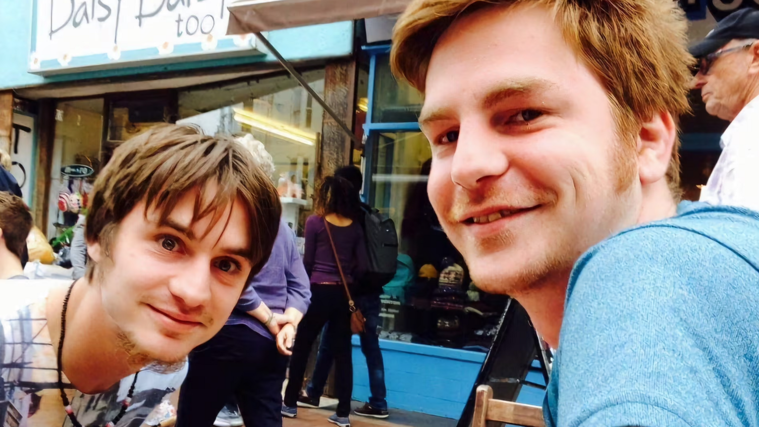 Two young men smile in a busy street outside some shops. 