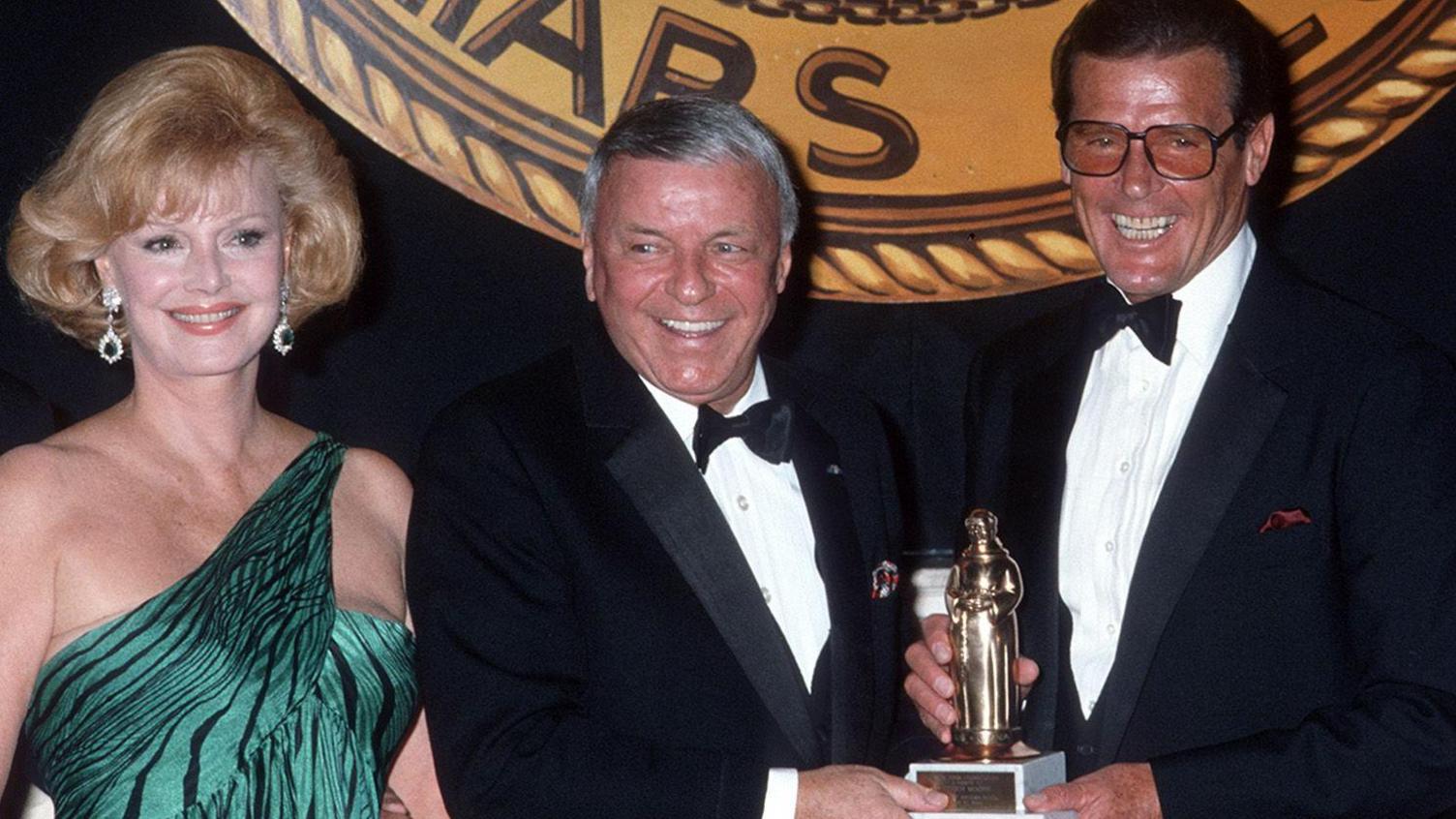 Sir Roger smiles as he poses for a picture with singer Frank Sinatra and his wife Barbara. Sir Roger and Frank Sinatra are holding a trophy together. They are dressed smartly in dinner jackets and bow ties. Sir Roger is wearing glasses and looks overjoyed. Frank Sinatra's wife Barbara is standing next to her husband. She has blonde hair, and is wearing a green, off-the-shoulder, evening dress with black tiger stripes on it. She has dangly emerald earrings.