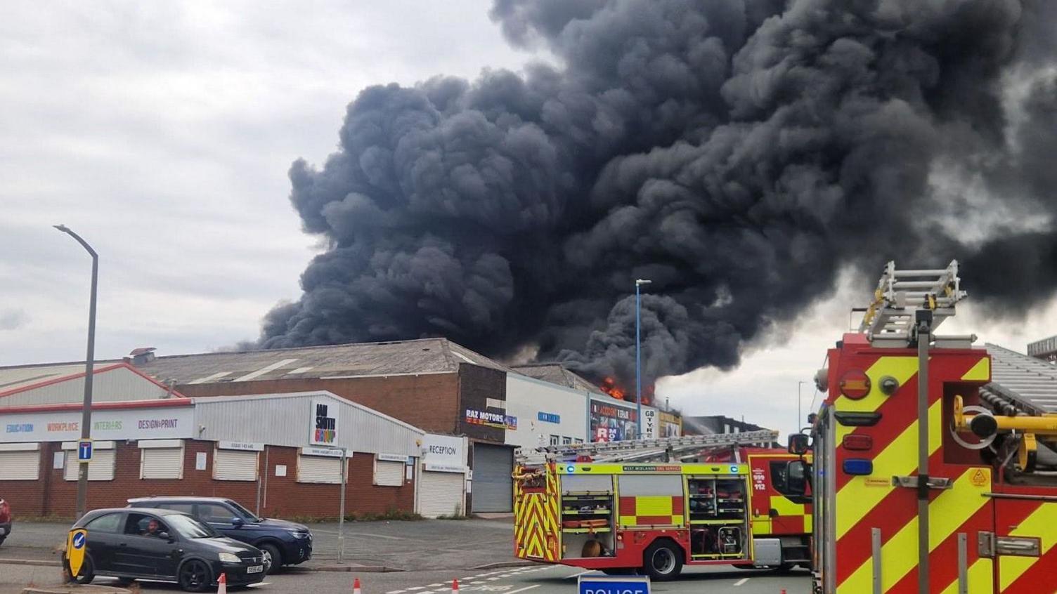 A factory fire has sent great plumes of smoke into the air. Two fire appliances can be seen outside it.