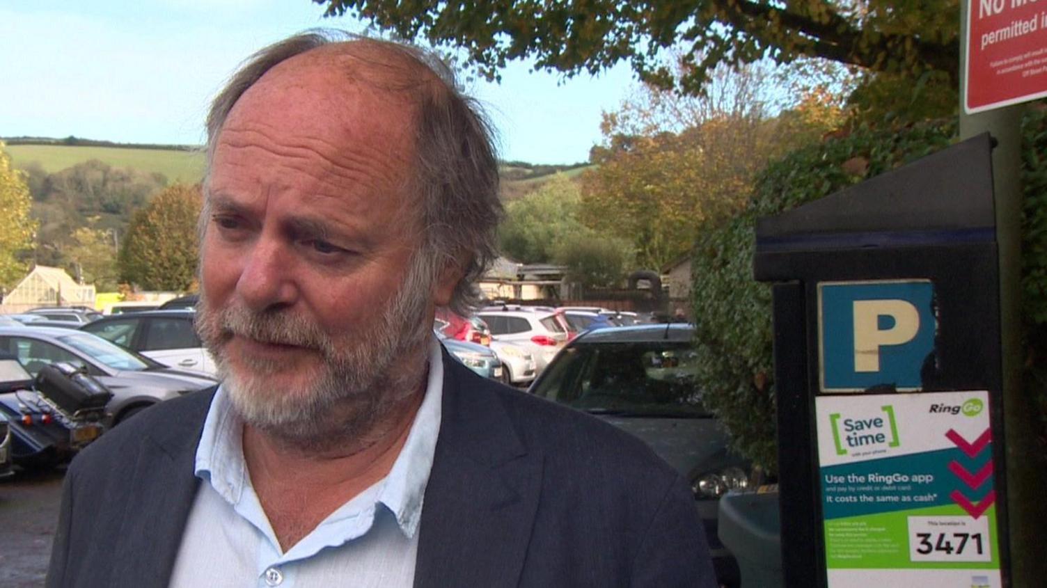 A man with a grey beard and a light blue open neck shirt and a dark jacket next to a parking machine in a car park.