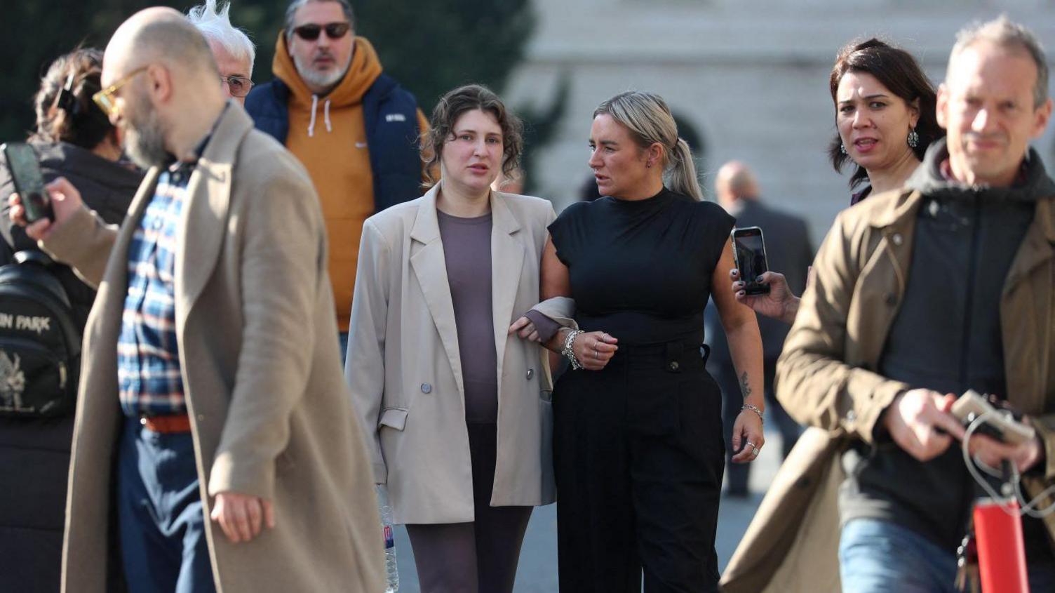 Bella Culley and her mother Lyanne Kennedy walk arm-in-arm through a group of people on a street in Tiblisi. Bella Culley is wearing a dark grey top and trousers with a light coloured formal jacket. Lyanne Kennedy, who has long blonde hair tied in a pony tail, wears a dark sleeveless top and trousers.
