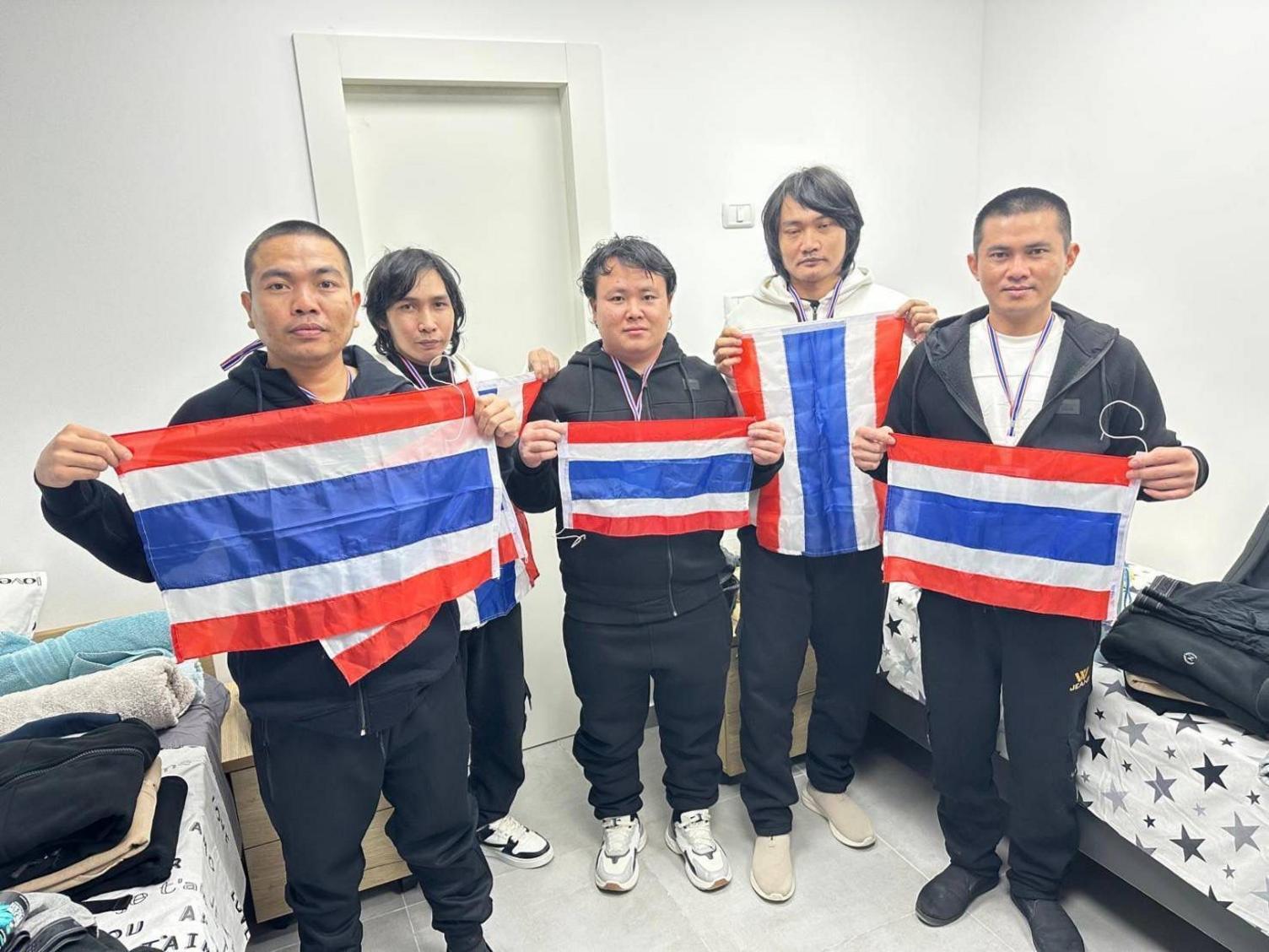 The five released hostages stand holding Thai flags in a white room. They are wearing tracksuits and trainers, with lanyards round their necks