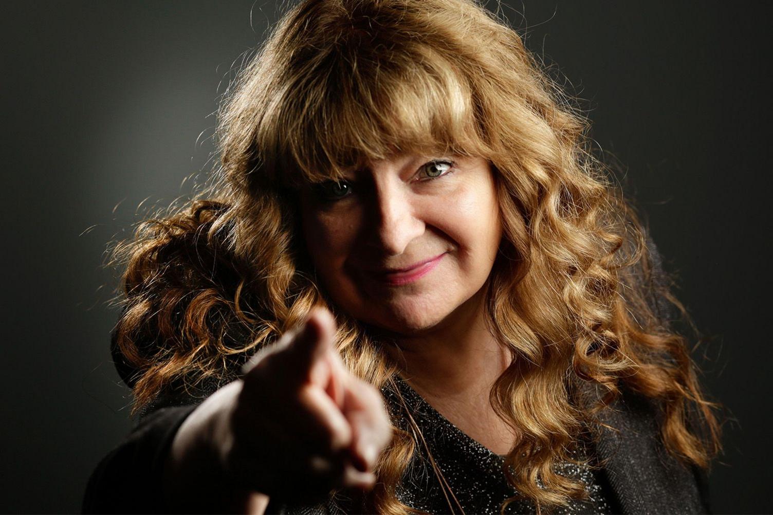 Publicity shot of Janey Godley pointing straight at the camera and smiling. She has long auburn hair, slightly curly and is wearing a black top with silver flecks