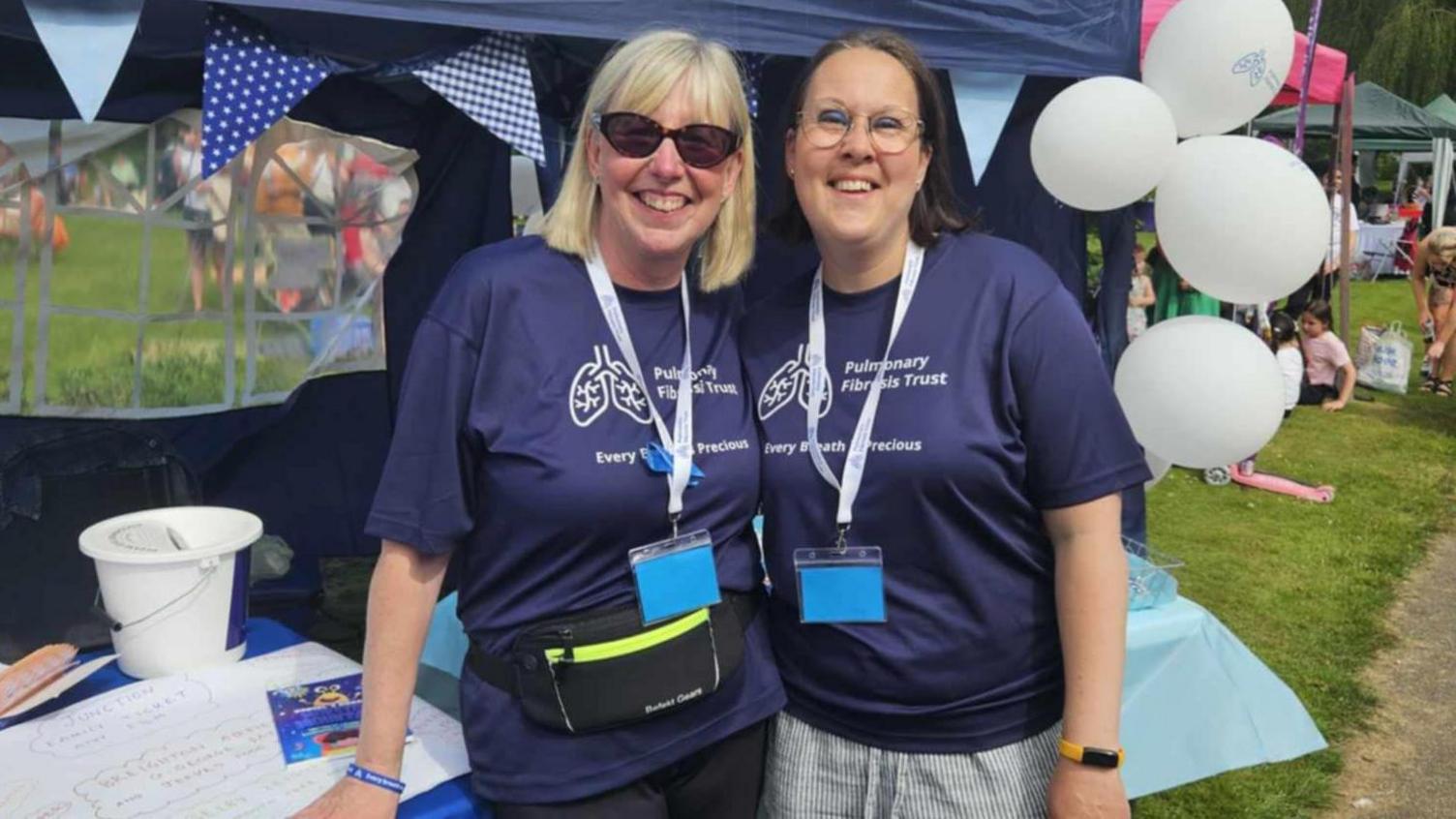 Joanne (left) and Nicola Lovejoy-Waud are stood next to a blue stall at an annual fundraising event for Pulmonary Fibrosis, called Picnic in the Park. They are both wearing navy blue T-shirts with the words "Pulmonary Fibrosis Trust, Every breath is precious".