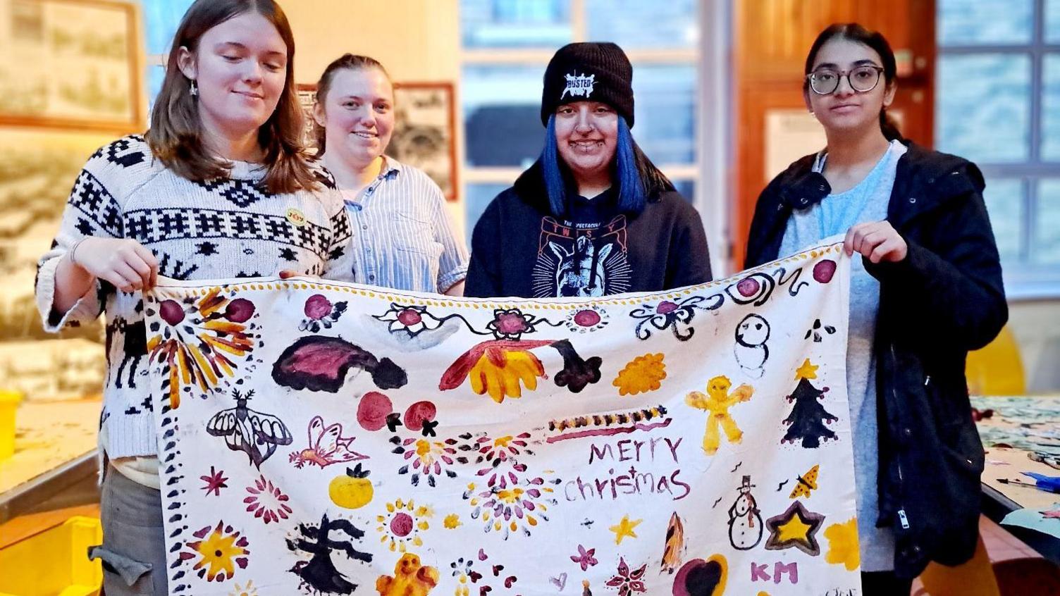 A group of four young women hold up a piece of cloth with yellow, black and pink drawings on it. It also says Merry Christmas.