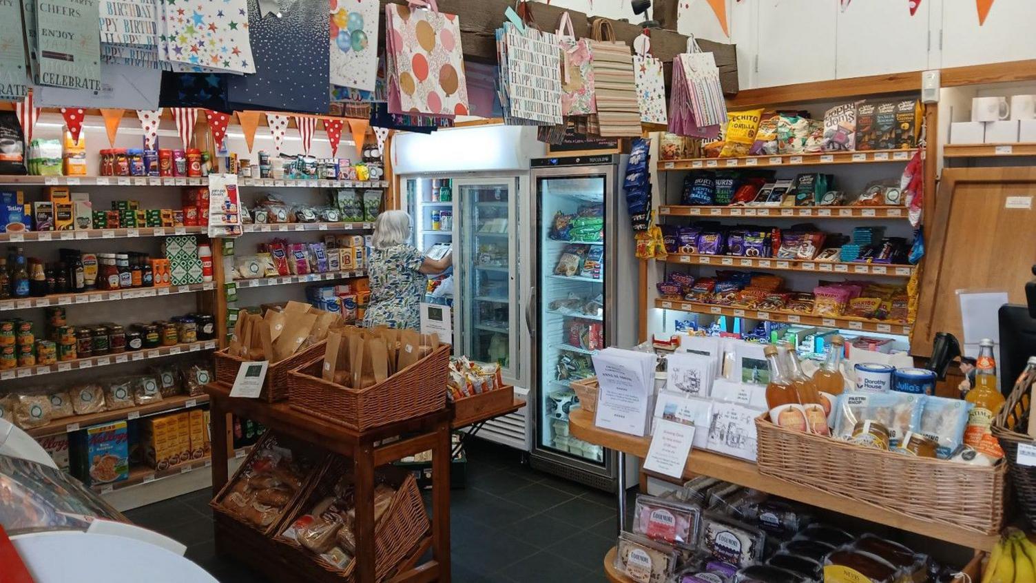 The inside of a community shop - there are shelves full of tins and jars. There are wicker baskets full of cider and honey and baked goods on the other side. At the back of the shop is a shelf with rows of chocolate. There are gift bags hanging from the ceiling. A woman is pictured getting something out of a fridge.