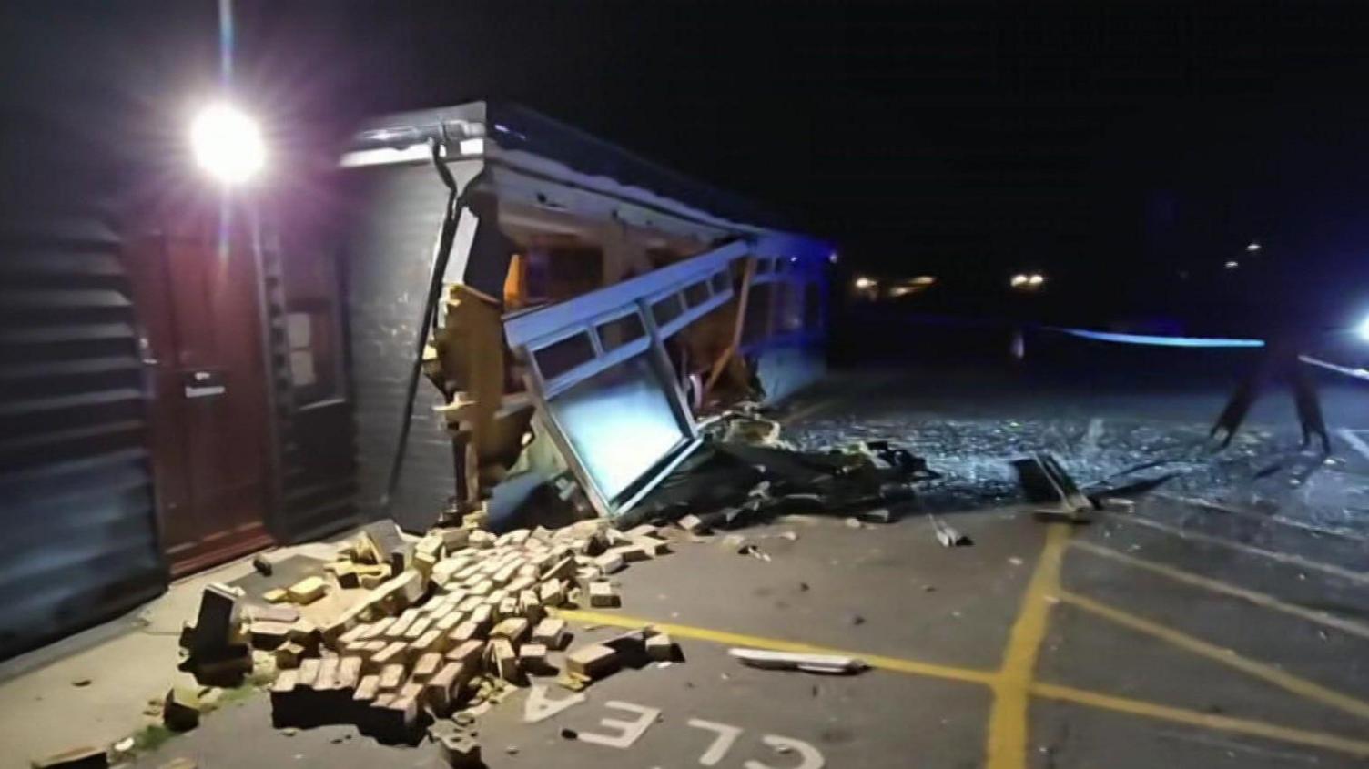 A black, single-storey building at the side of a tarmacked area, at night. There is broken glass and several bricks strewn across the tarmac and a big hole in the side of the building. White guttering and windows are hanging off the damaged building facade.