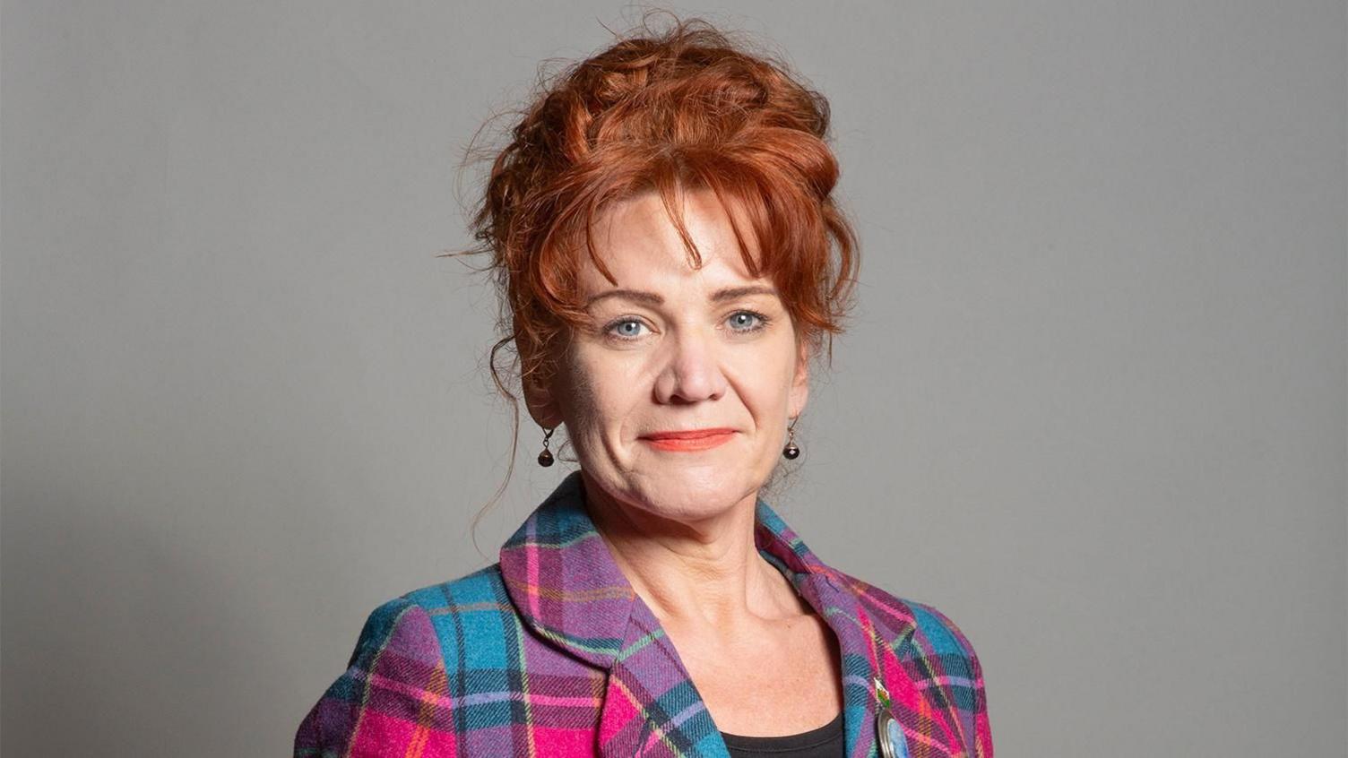 A headshot of Sarah Atherton. She has ginger curly hair which is tied back, a fringe, and blue eyes. She is wearing coral lipstick, round drop earrings and a pink and blue plaid jacket. She is standing in front of a grey background.