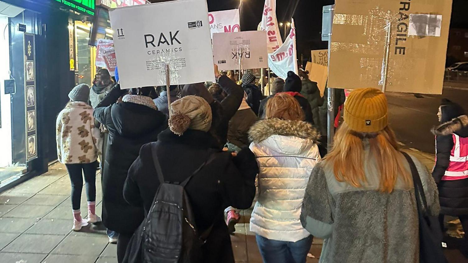 A back view of mostly women walking up a street in Northampton in 2024. They are holding up placards and are dressed for winter conditions with hats and coats 