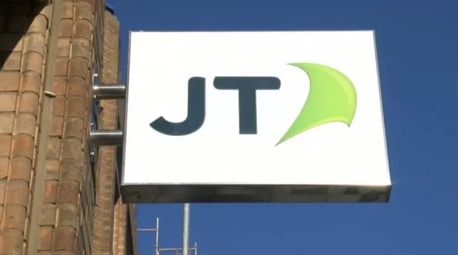 A brick wall to the left holds a metal sign on which is written JT on a white background with a green insignia, the sky in the background is blue with no clouds.