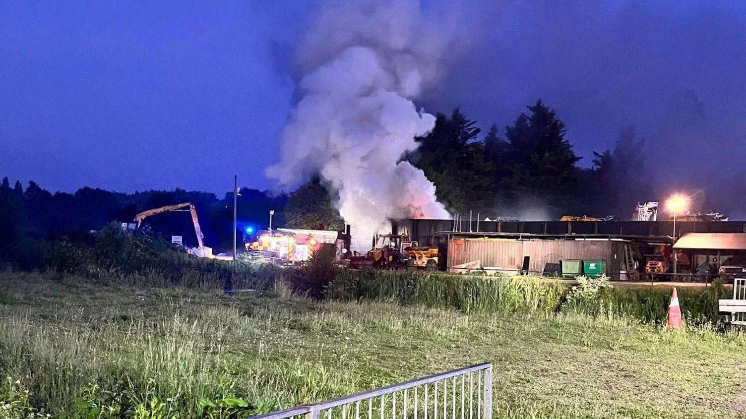 A fire at a metal recycling centre at Kings Langley. Smoke is rising above a low-lying building. To its left can be seen a fire engine. The sky above is the dark blue of night. In front of the building is an expanse of grass.