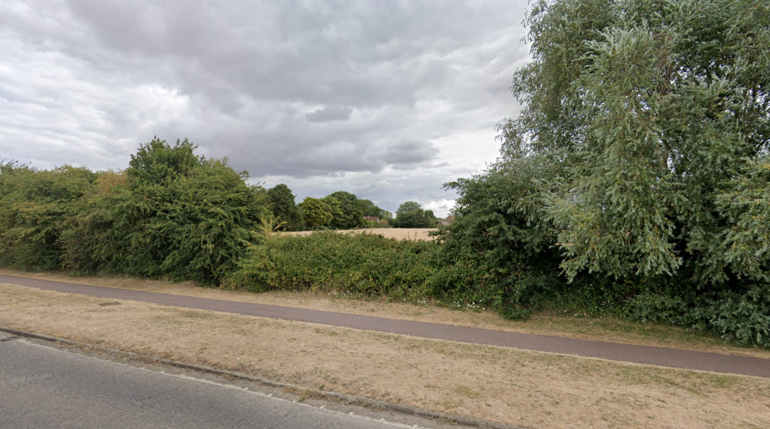 Street view image looking from the main road towards the fields that could have been built on. There are bushes separating the road and field and clouds in the sky. The land is flat and surrounded by trees. 