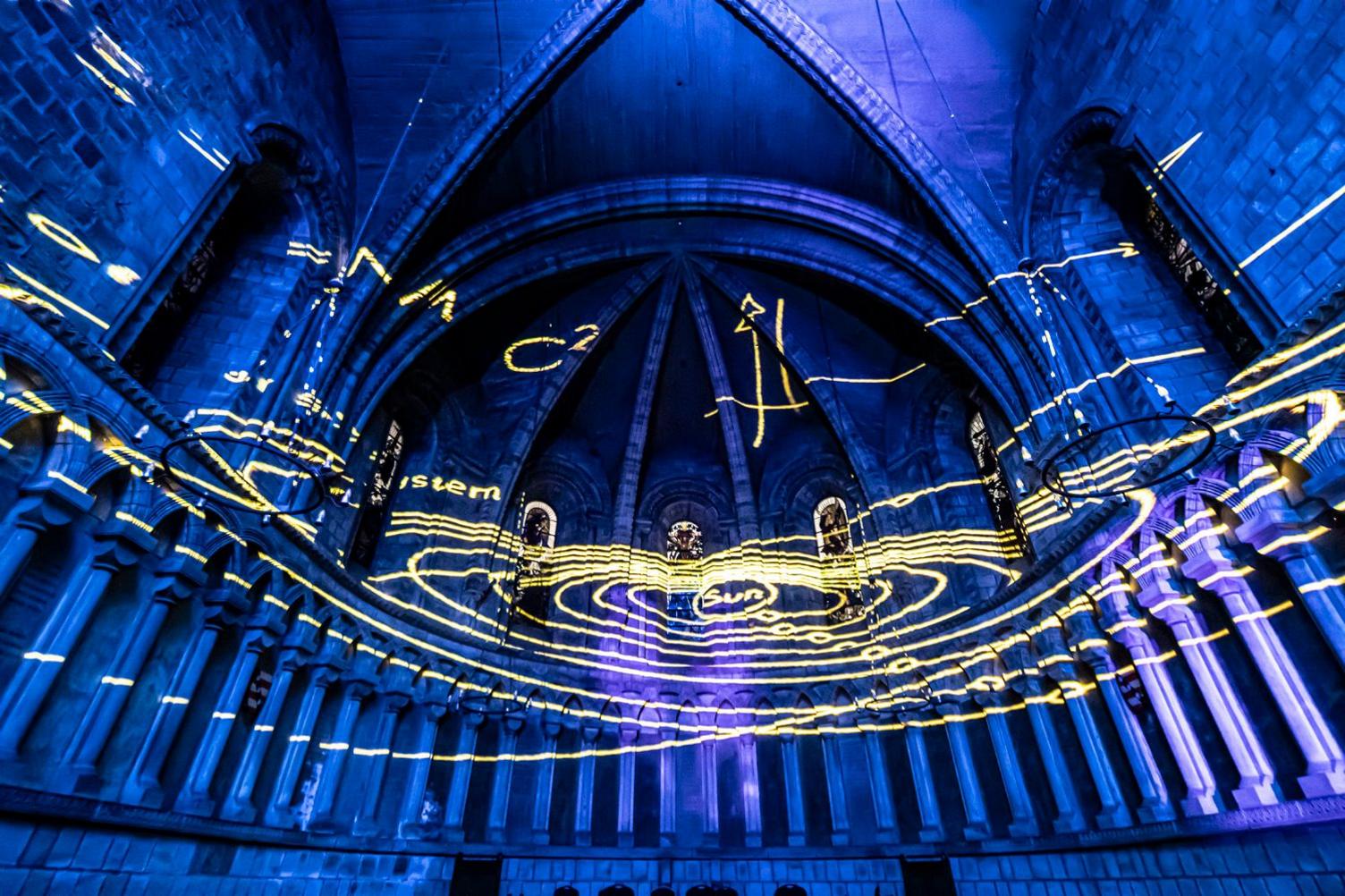 Intertwining yellow circles projected inside the cathedral against a blue background. Sun and system is also written in yellow. 