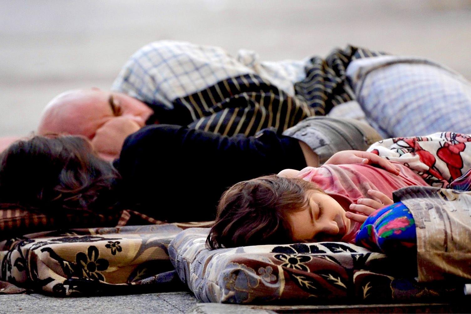 Father and  two children sleep outside on thin bedding.