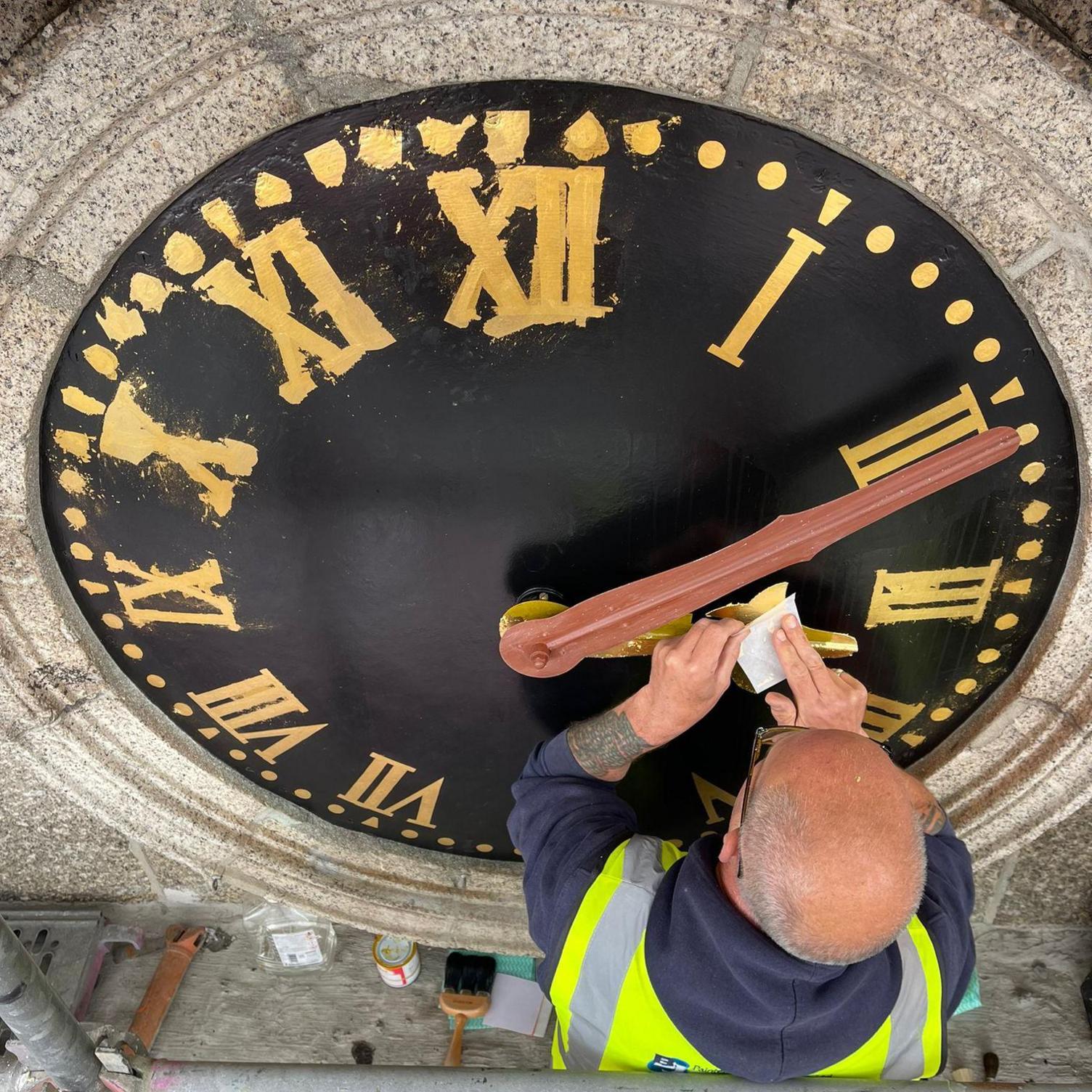 A person in a dark blue hooded top with a high vis vest over the top works on the clock. The clock has a black face and gold coloured detailing is being applied to one of the two hands