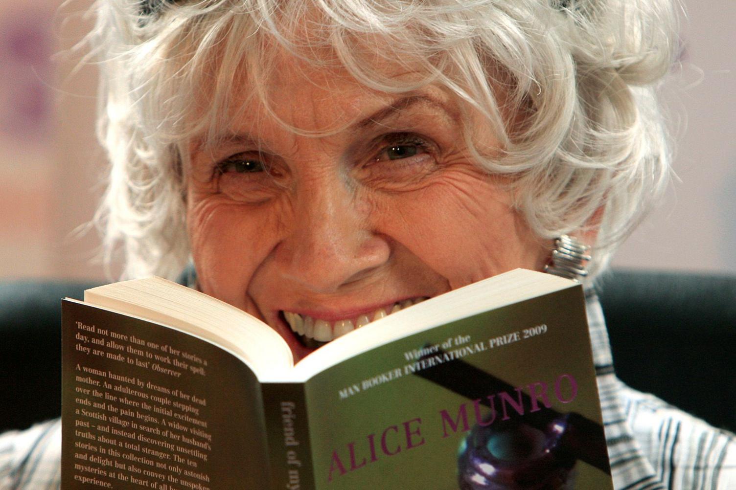 Alice Munro is photographed close up and laughing, holding one of her own paperbacks open in front of her. She has short grey slightly wild hair. 