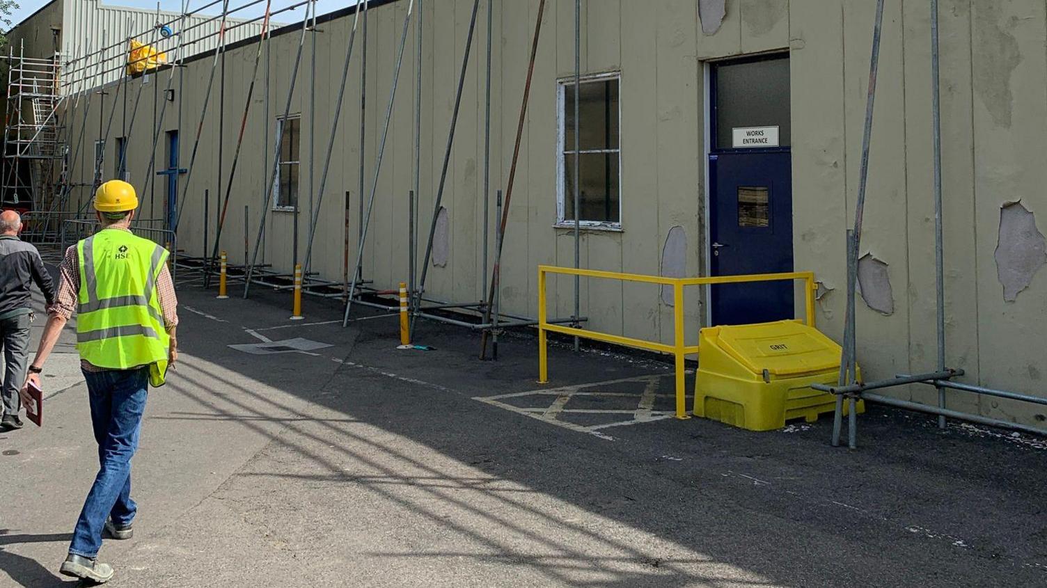 A worker in a hi-vis vest and hard hat walks in front of a factory building which is covered in scaffolding.