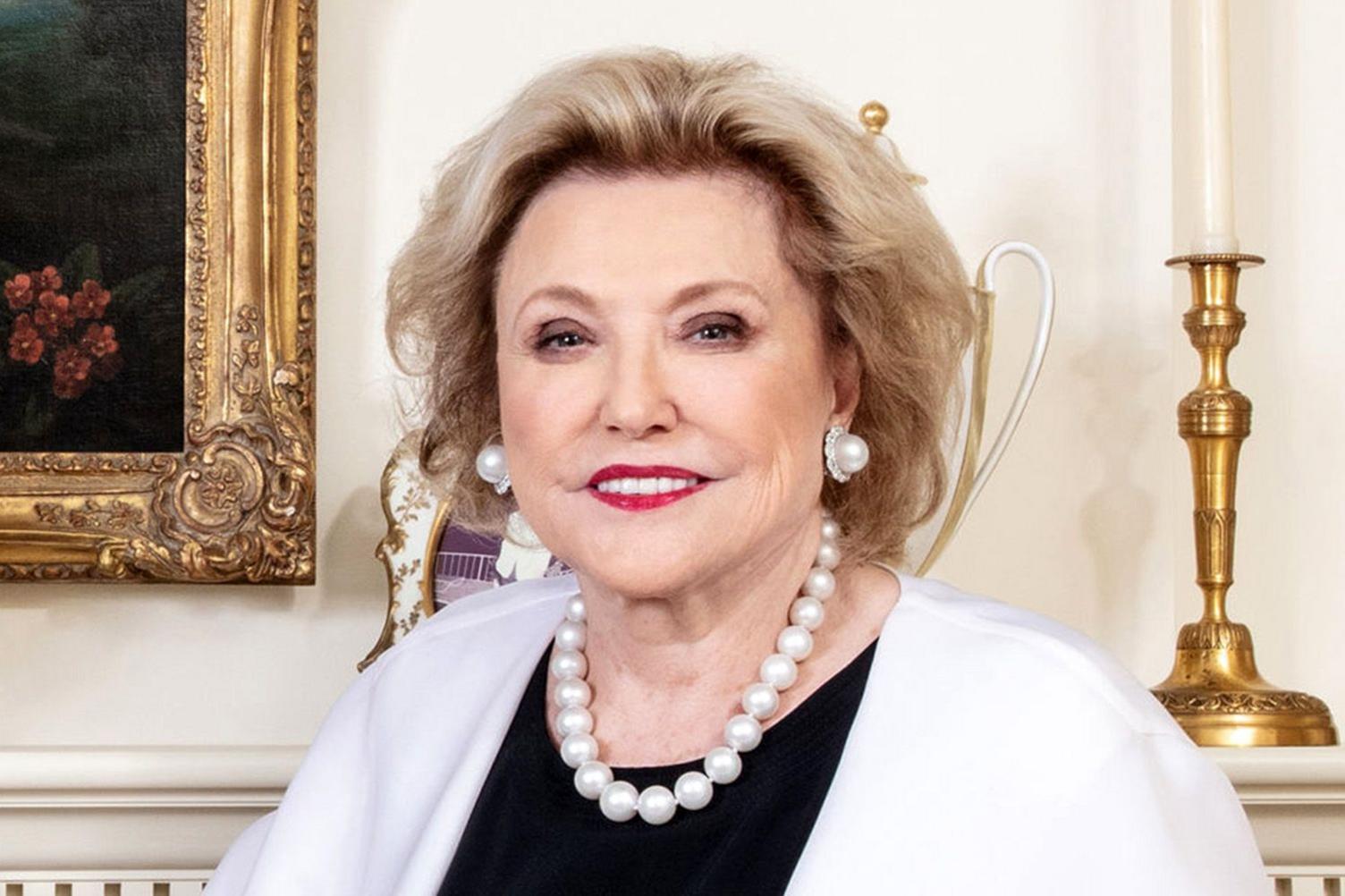 Barbara Taylor Bradford is photographed against what looks like an opulent fireplace. She has blonde hair and her smile reveals very white teeth. She is wearing a cream jacket over a black top and has matching pearl earrings and necklace.