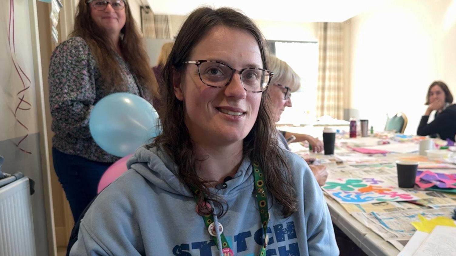 A picture of a woman smiling with brunette hair and glasses. She is sat by a table which is covered in arts and crafts