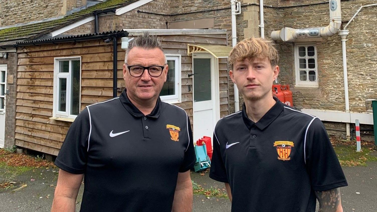 Two men standing in front of a building. The man on the left is Melvyn Wilkes, he has grey hair that is spiked up, and is wearing black glasses and a black football shirt. On his right is Kyle Wilkes, who has long blonde hair and is also wearing a black football shirt