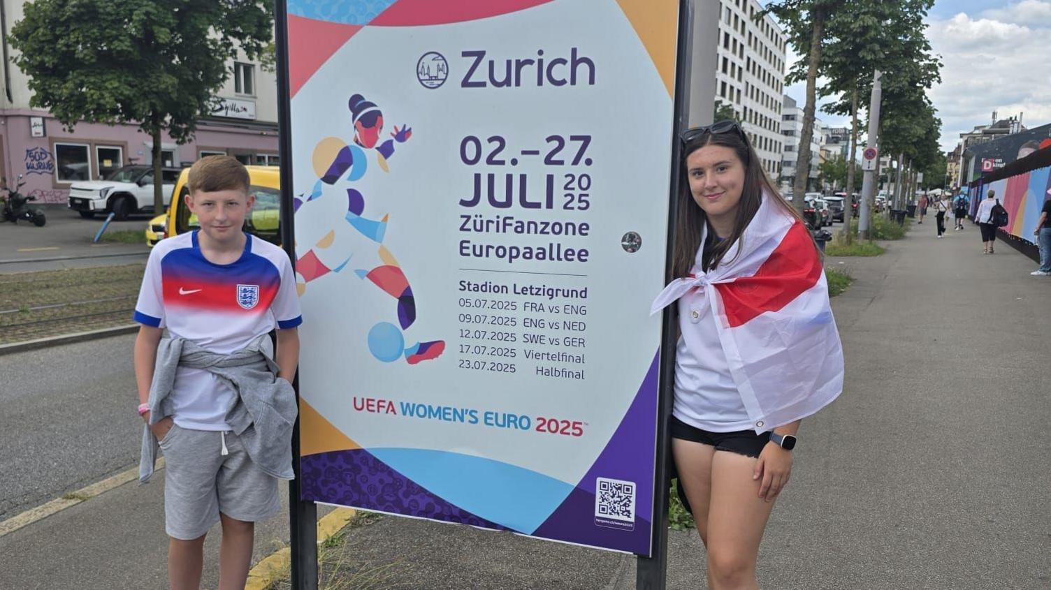 Ethan and Amelia stand next to a sign advertising dates for the games. Amelia is draped in an England flag.