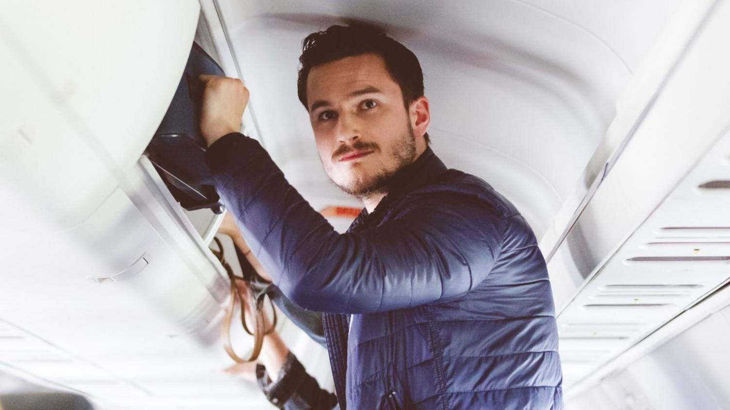 Man standing up on a plane with hand on luggage in the overhead compartment