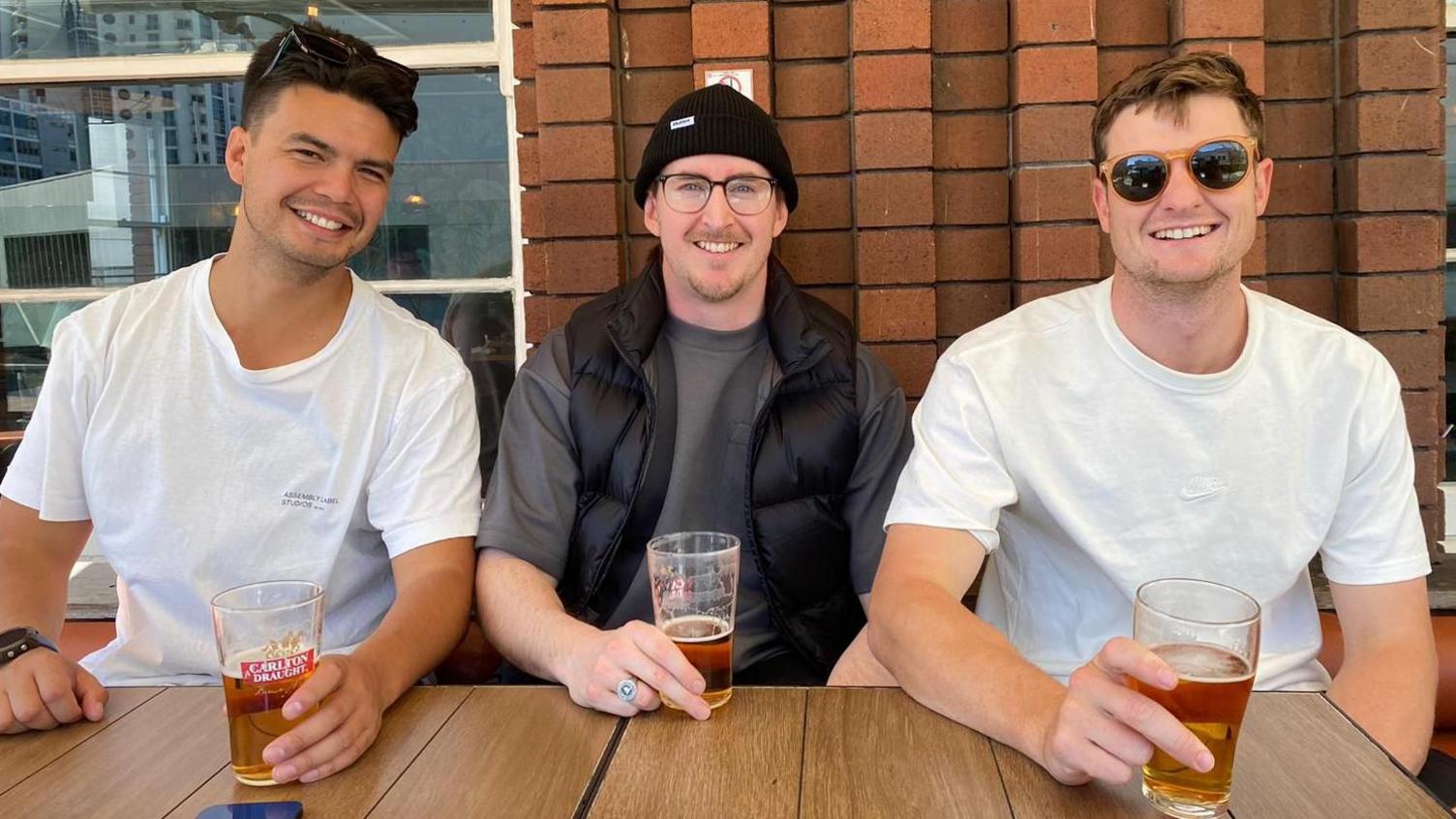 three young men sitting at a table, each holding a pint of beer and smiling at the camera