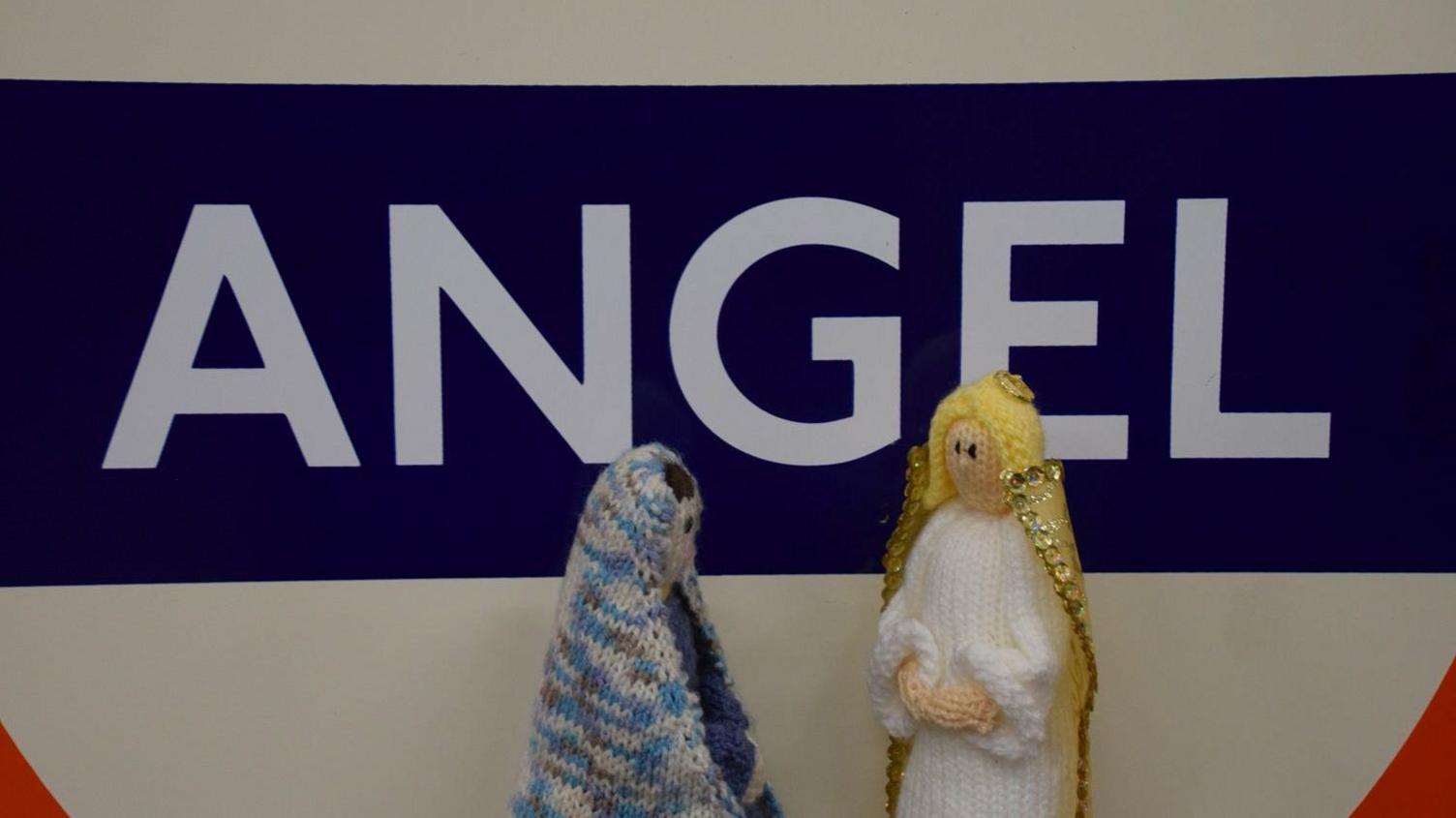 Miniature knitted figurines of Mary and angel Gabriel in front of an Angel station sign