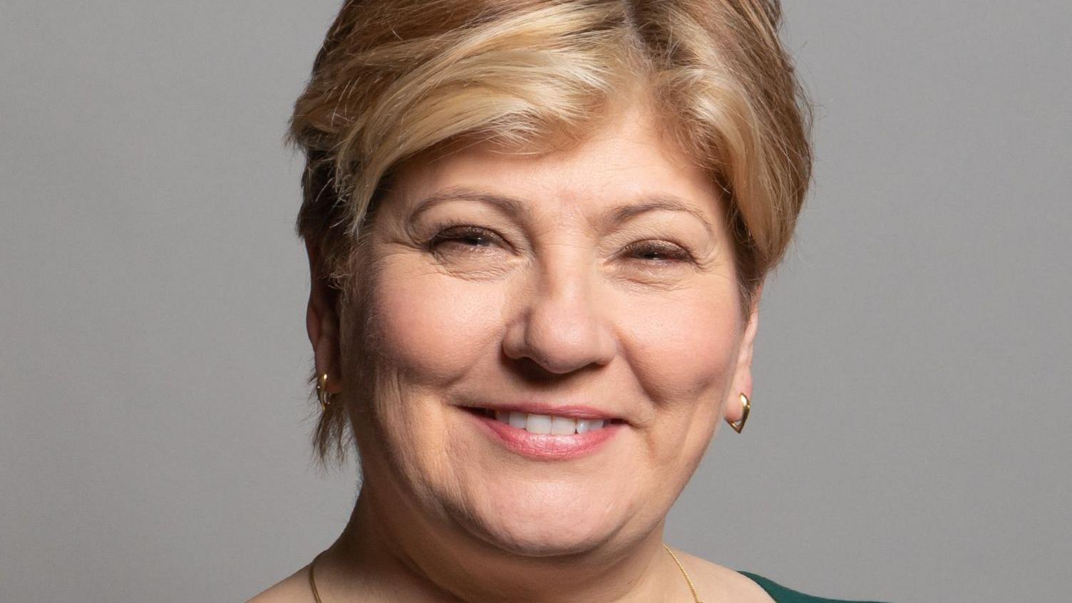 Dame Emily Thornberry in an official photograph. She is wearing a green dress with a square neck and a slit at the front, gold earrings and necklace, and she is smiling at the camera.