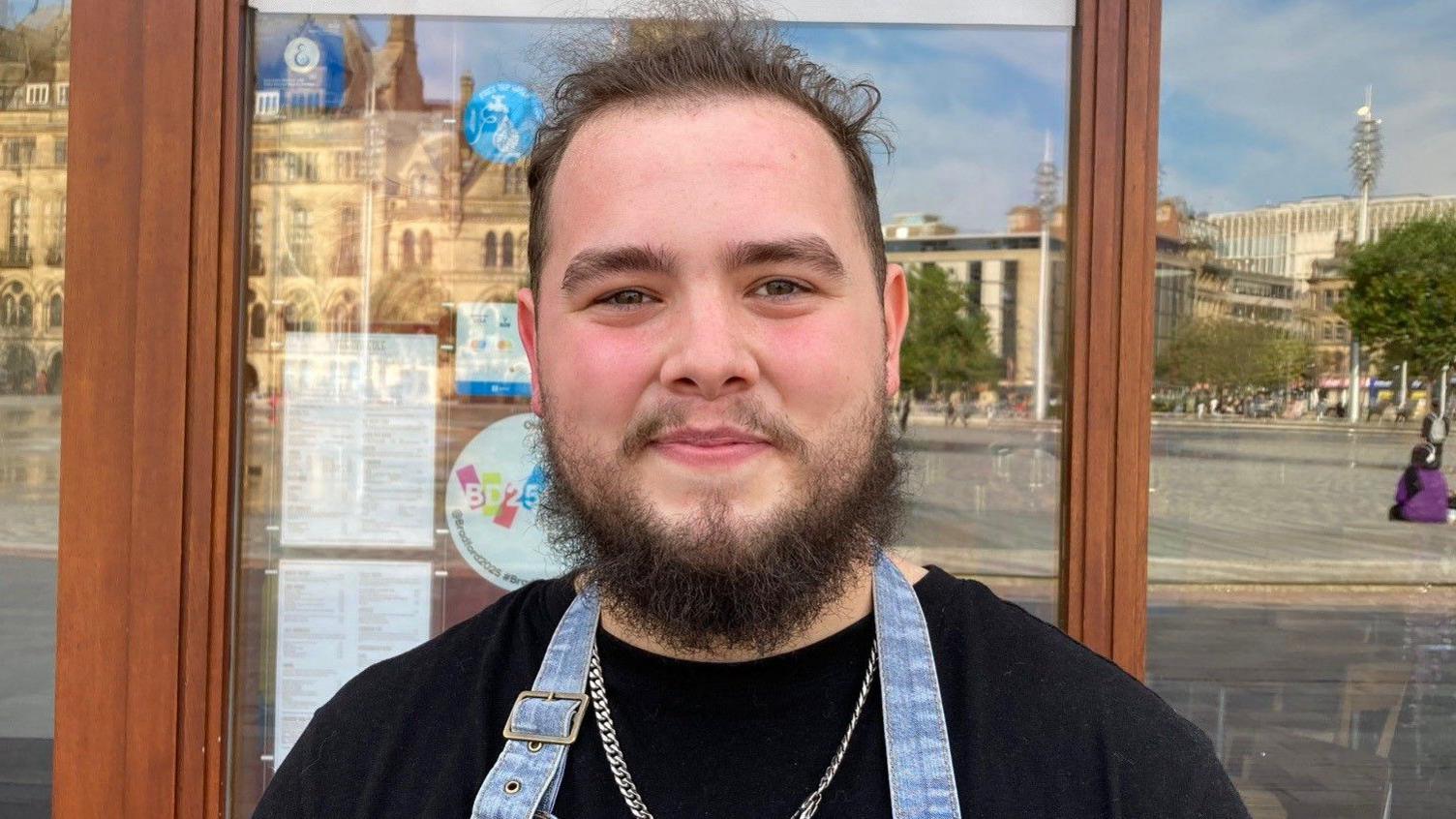 A young, white male with a dark brown beard and fuzzy brown hair stands in front of a cafe. He is wearing a black t-shirt and apron