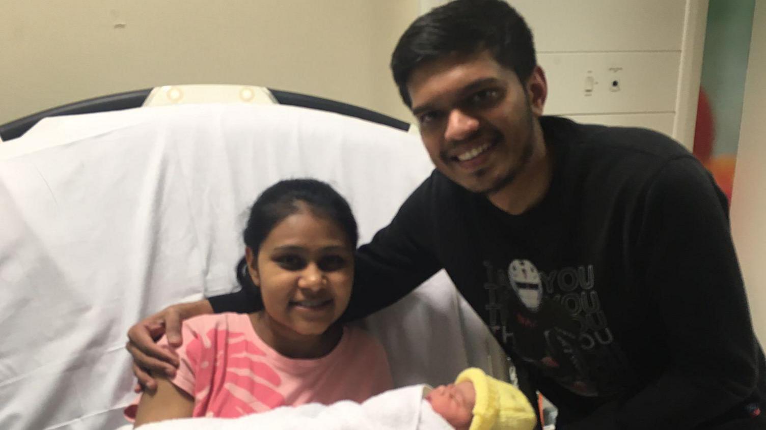 An Indian couple carrying their newborn daughter, who is wearing a yellow knitted hat, posing for a picture at Leicester Glenfield Hospital