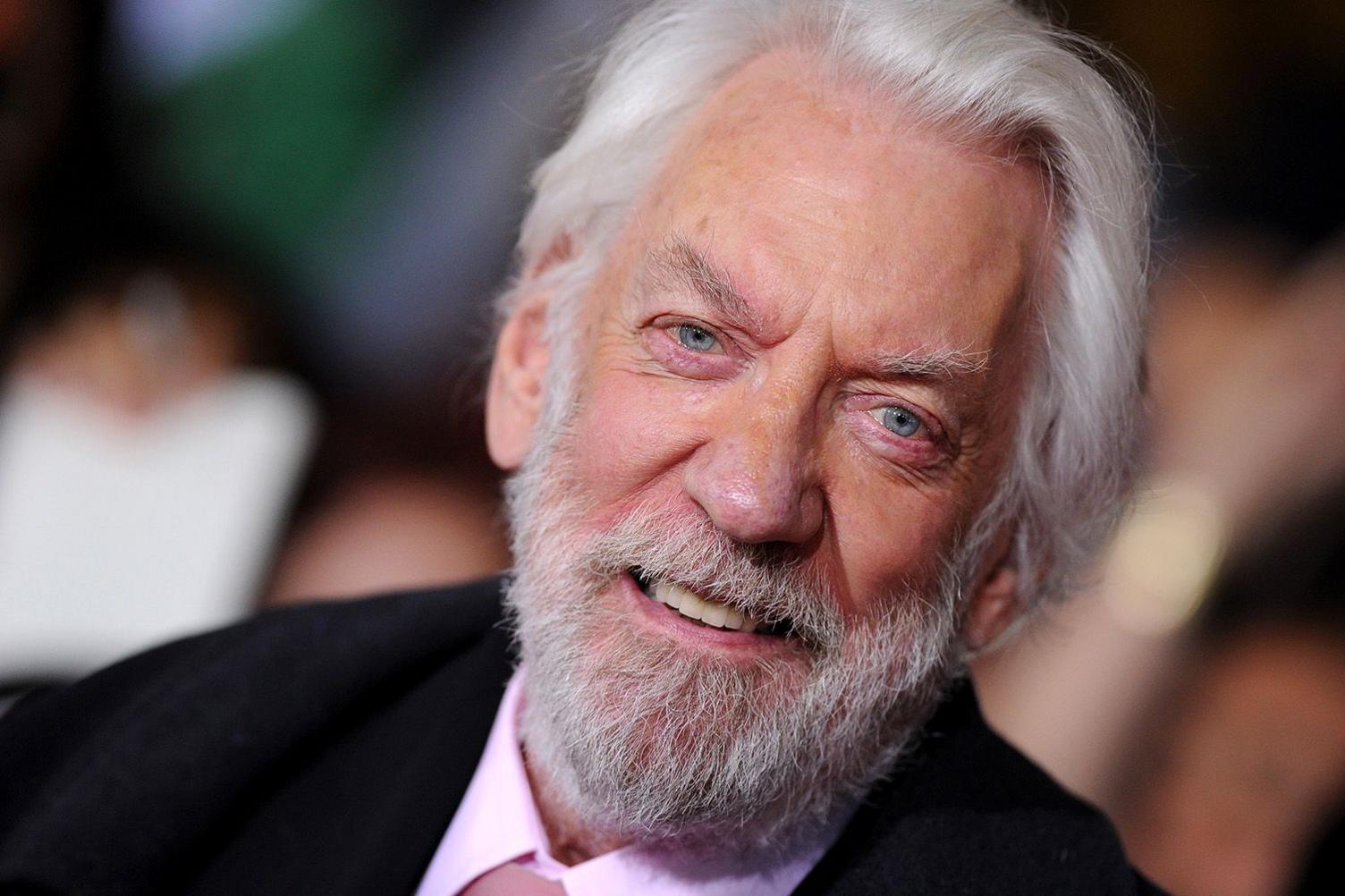 Close-up photo of Donald Sutherland pictured at the premiere of The Hunger Games: Catching Fire in 2013, when he was 78. He is grey-haired and bearded and is smiling. He wears a black jacket and a pink shirt and tie.