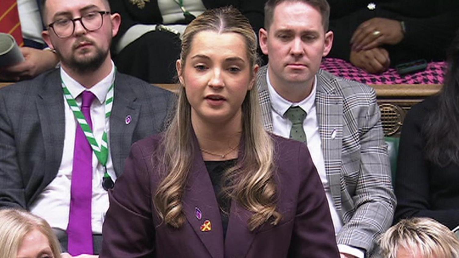 A woman in a lavender blazer stands up in front of two men in suits in the House of Commons.
