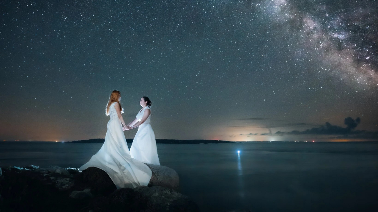 Two brides hold each other's hands while standing on a rock near the sea with an island behind, beneath the Milky Way and hundreds of shining stars.