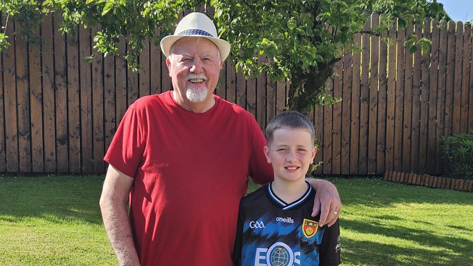 An elderly man with a straw cream hat, white goatie beard and red t-shirt on smiling in his garden and putting his arm around his a boy who is up to his shoulders who is wearing a black and blue o'neills shirt and has brown short hair and is smiling. The garden is small and green with a brown fence. 