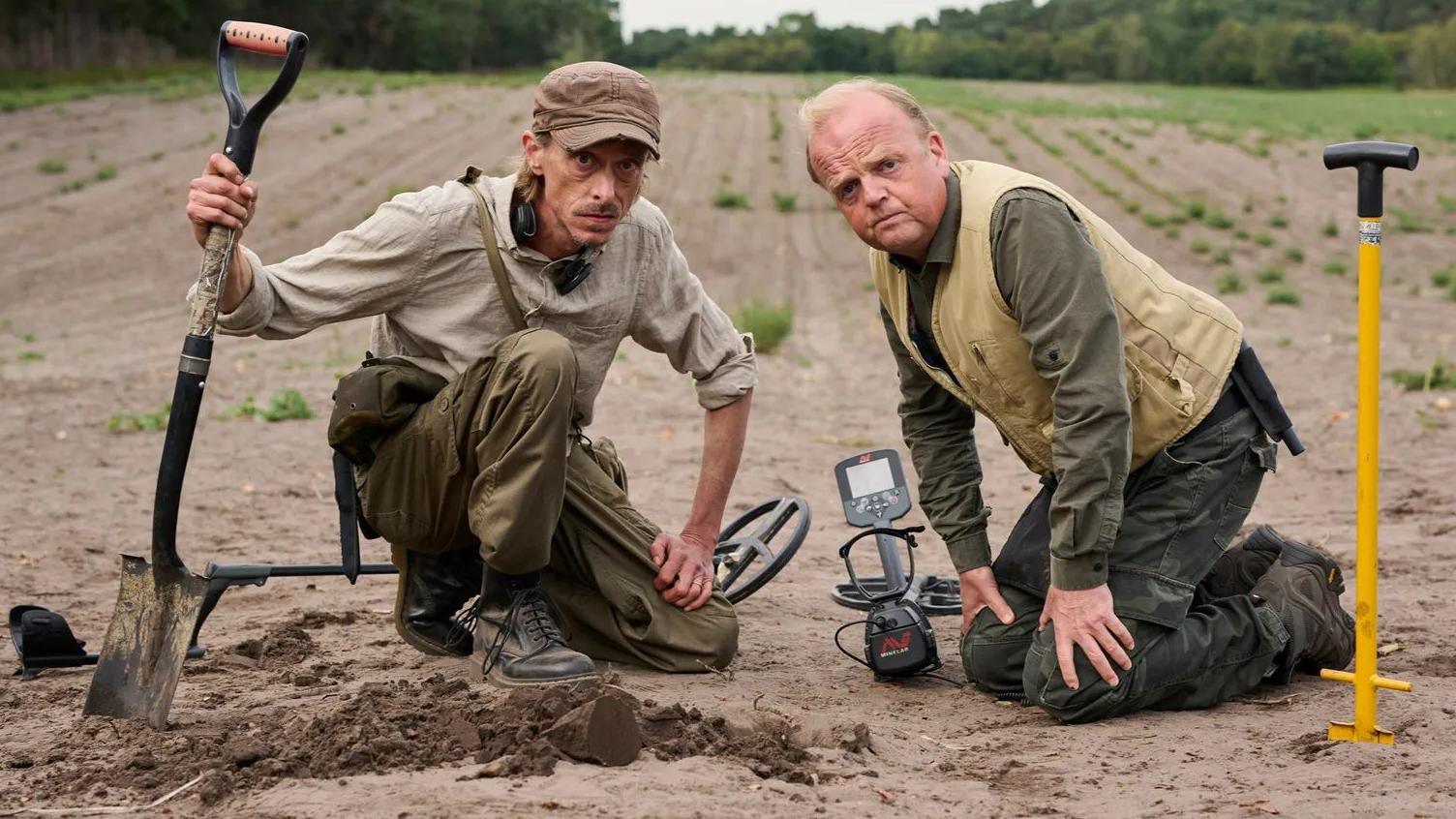 The two main actors in Detectorists Mackenzie Crook and Toby Jones pose in character kneeling on a field with their tools. Crook holds a spade and next to Jones is a yellow digging tool and black detecting kit. They wear boots and outdoor, khaki coloured clothes.