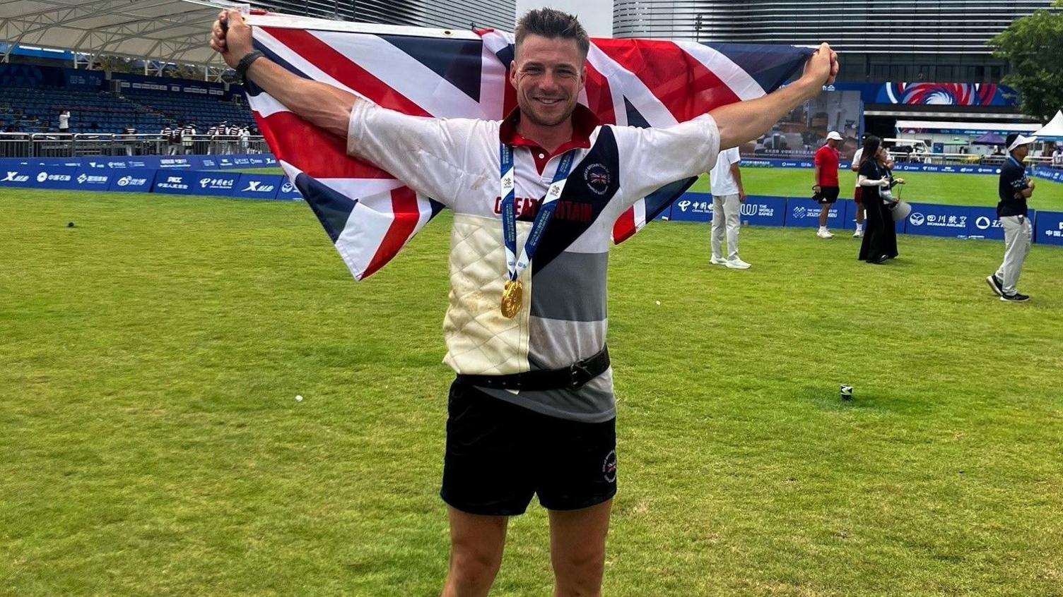 Joe Birch is standing on a sports pitch wearing his gold medal and holding a British flag outstretched behind his arms. He is smiling broadly. 