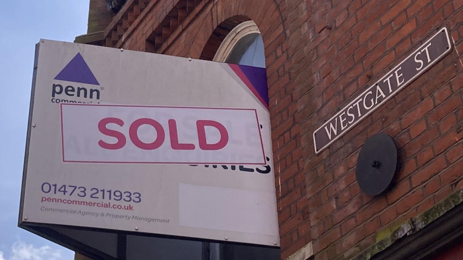 A sold sign hangs from a red brick building window. A street sign that reads Westgate Street has been placed on the building next to the sign.