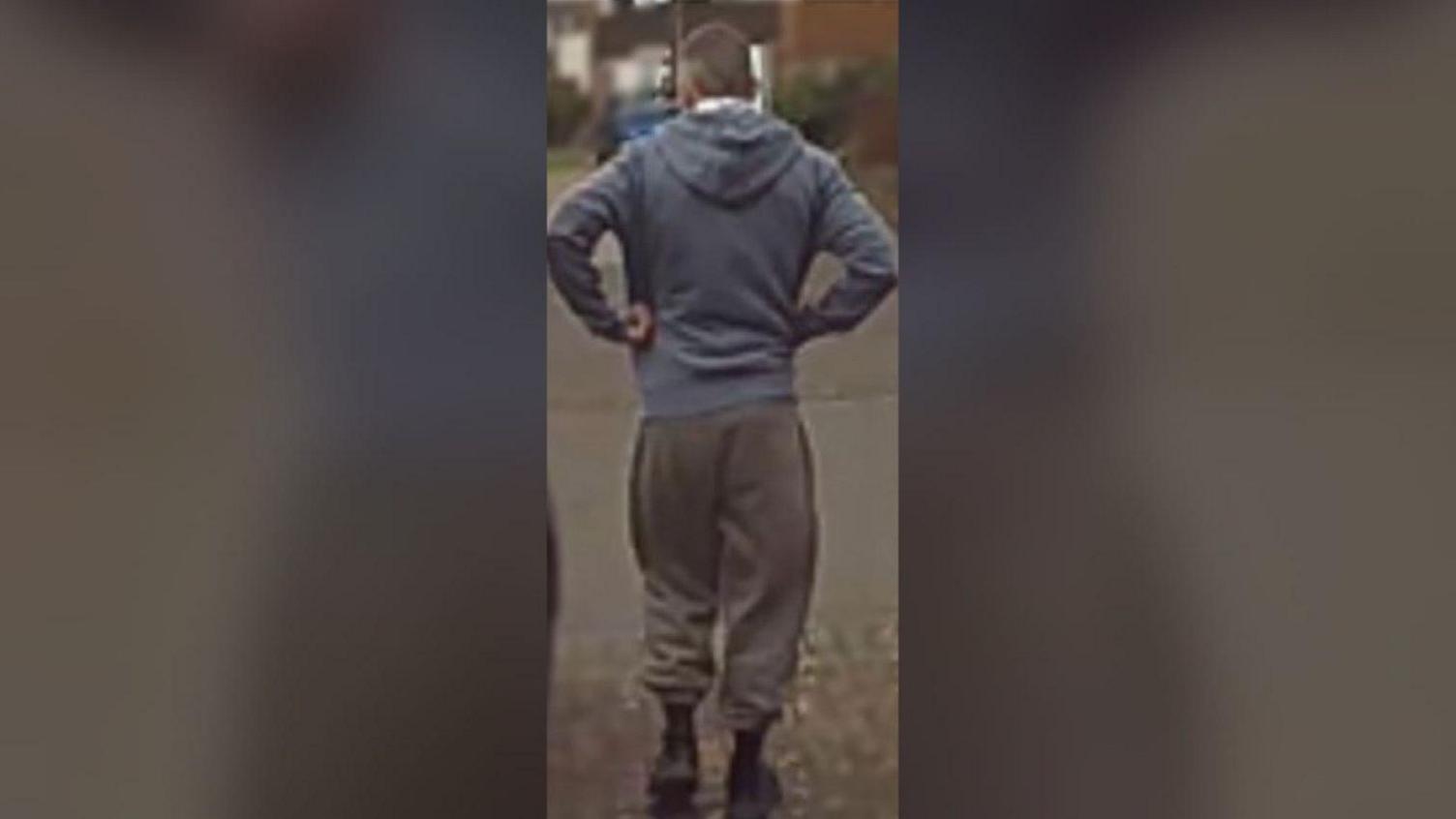 The back of James on a CCTV camera, wearing brown tracksuit bottoms, a grey zipped hooded top, a white shirt with a light brown and blue chequered pattern, black socks and black material trainers